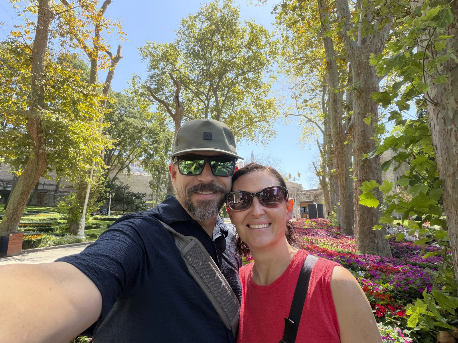 A couple in sunglasses taking a selfie under plane trees, with pink flowers and a stone wall behind them.