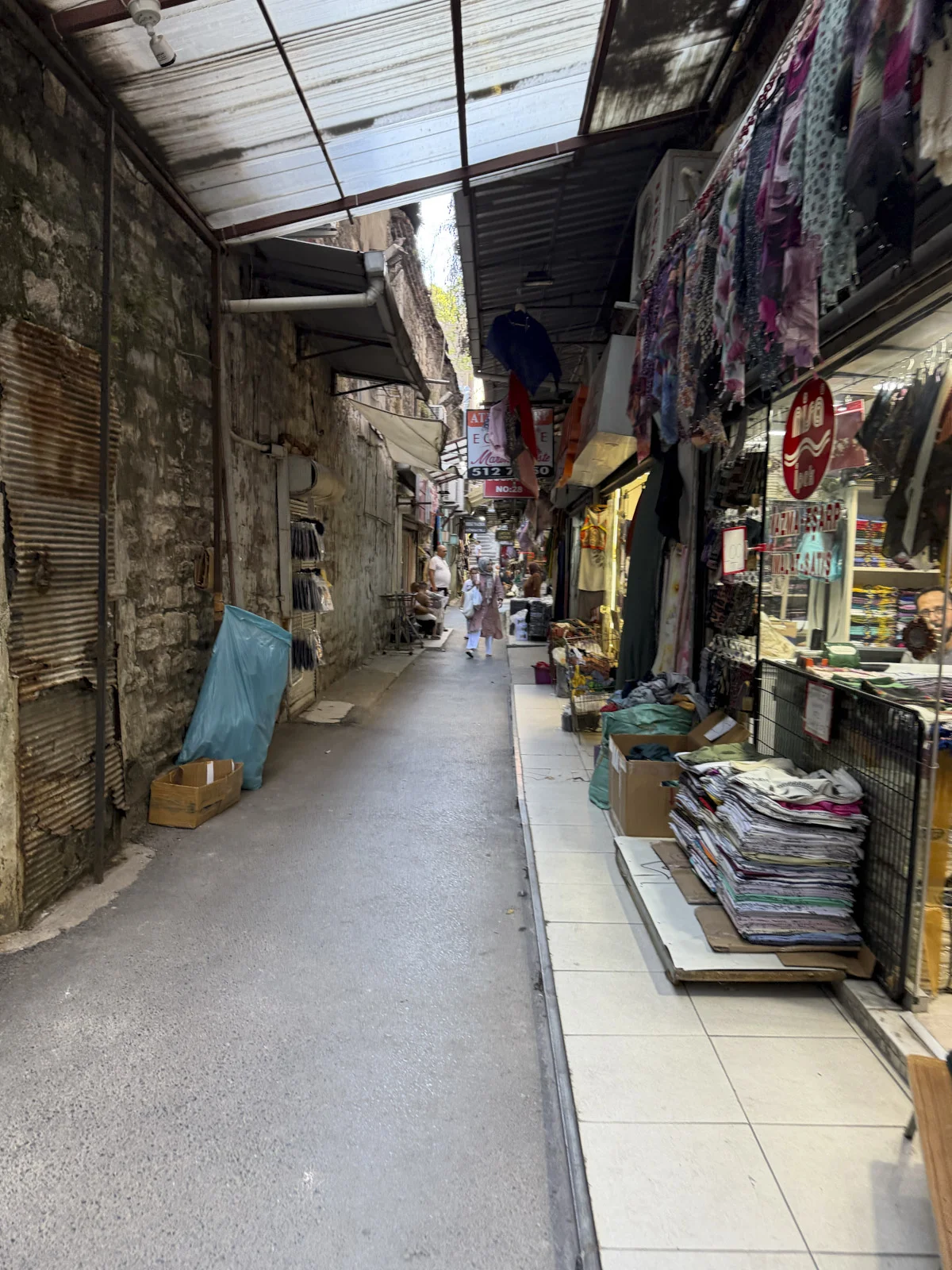 A narrow covered market alley with hanging clothes, stacked fabric, and a rough stone wall.