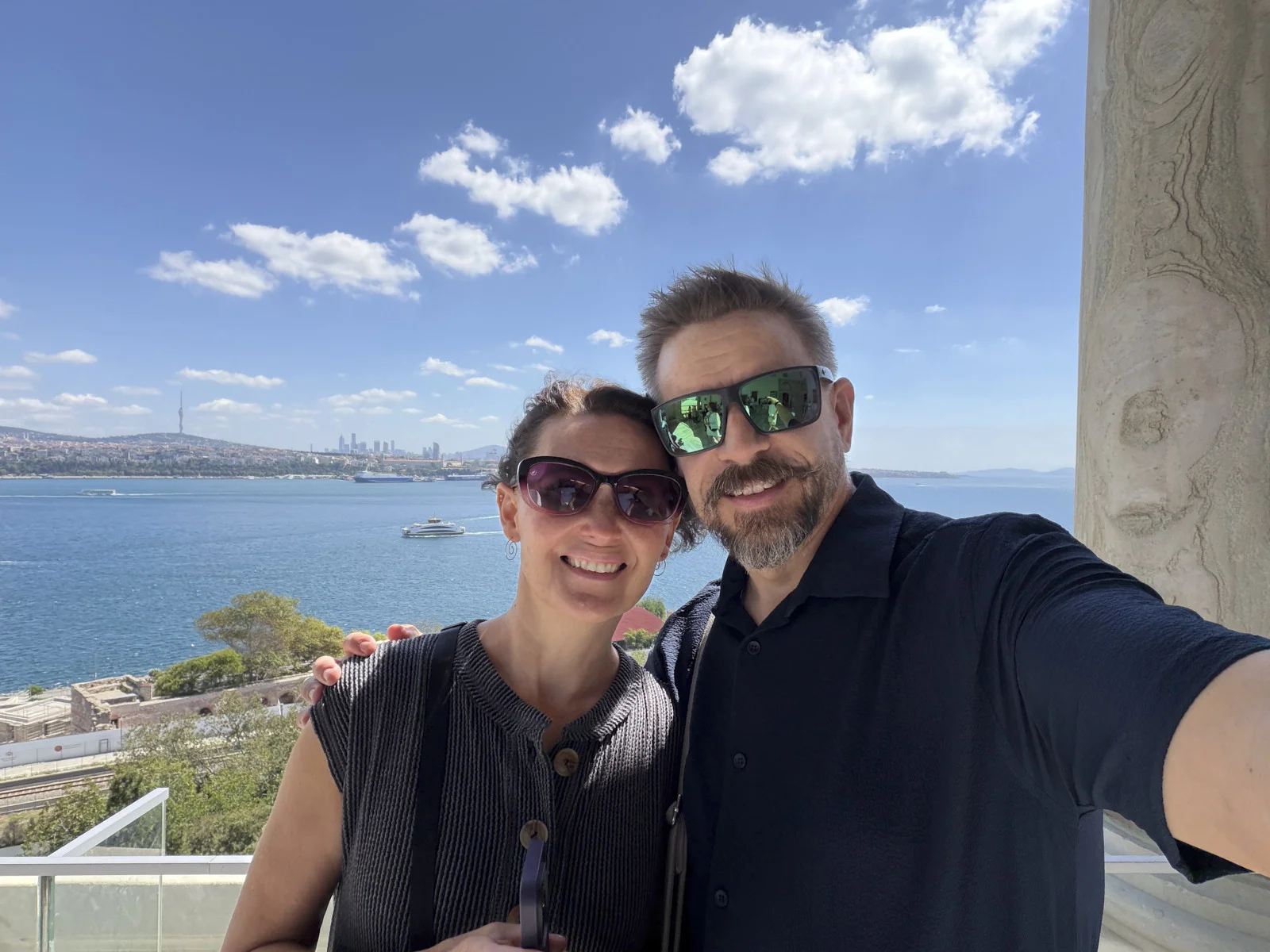 A couple taking a selfie on a stone terrace above blue water, with a distant city skyline and a boat.