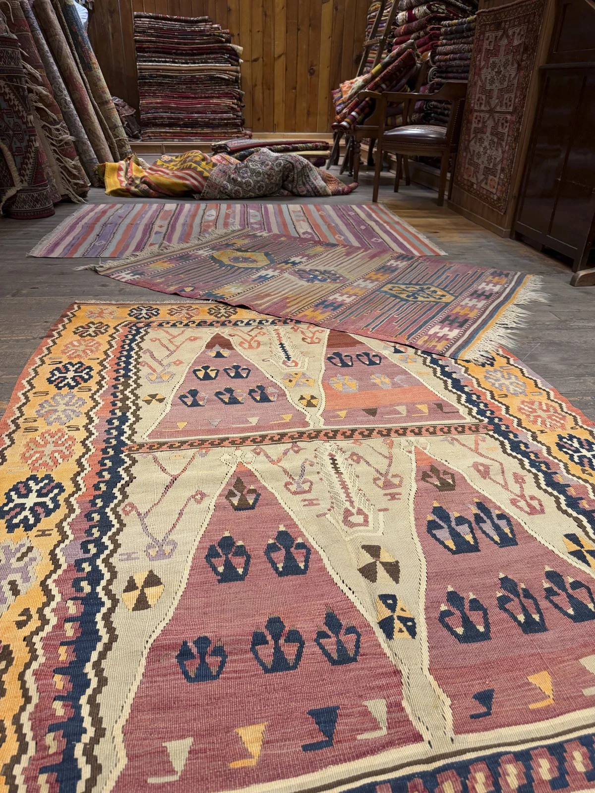 Large geometric kilims spread on a wooden floor in a rug shop, with more rugs stacked along the wall.