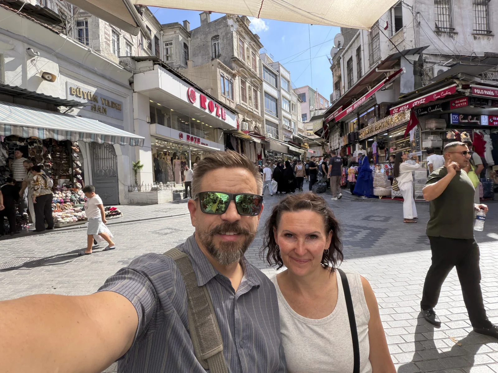 A couple taking a selfie on a busy market street, with the Büyük Valide Han sign and shop awnings behind them.