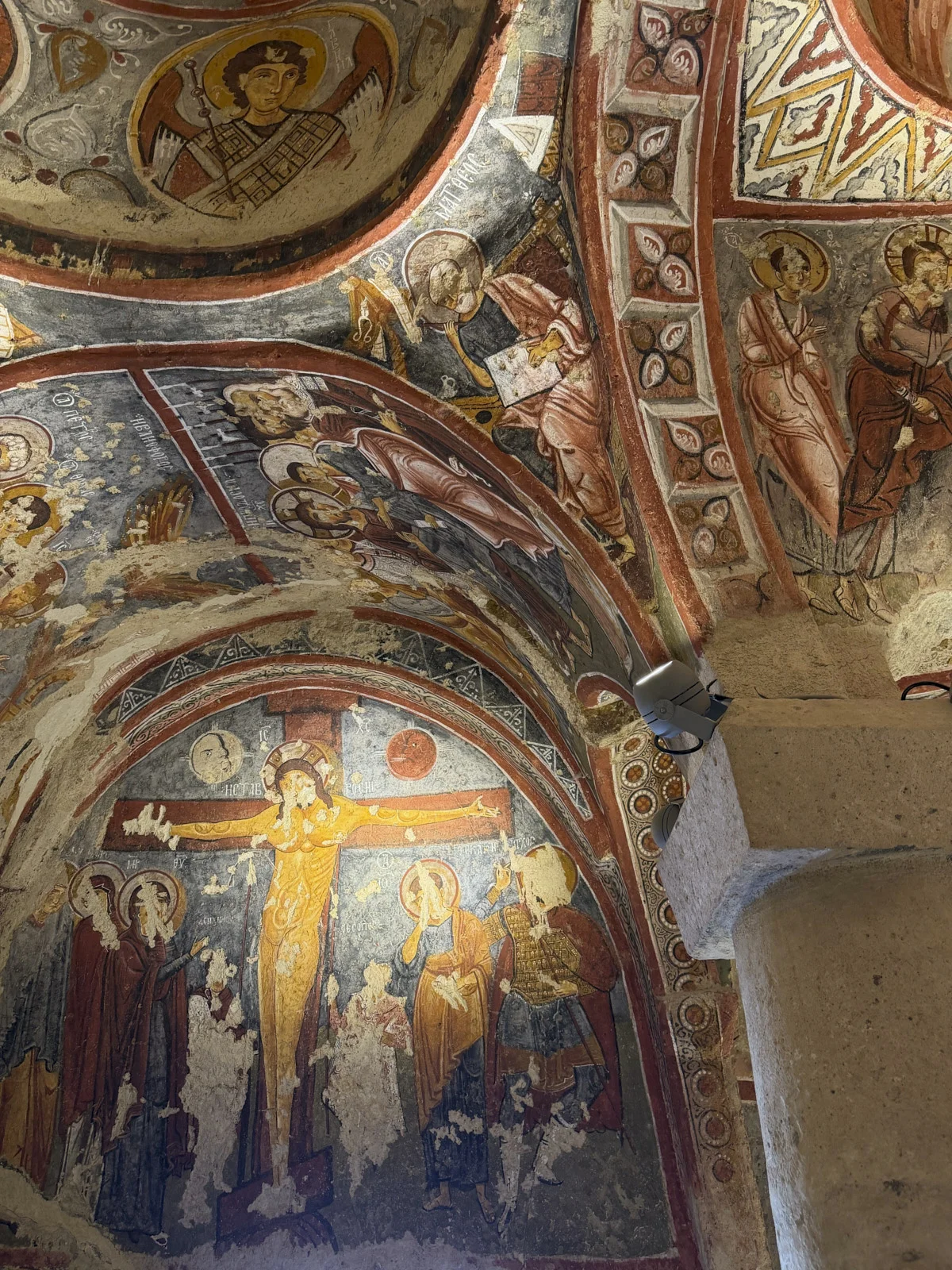 A worn cave-church fresco of the Crucifixion in yellow and red, with saints and geometric borders on the vault.