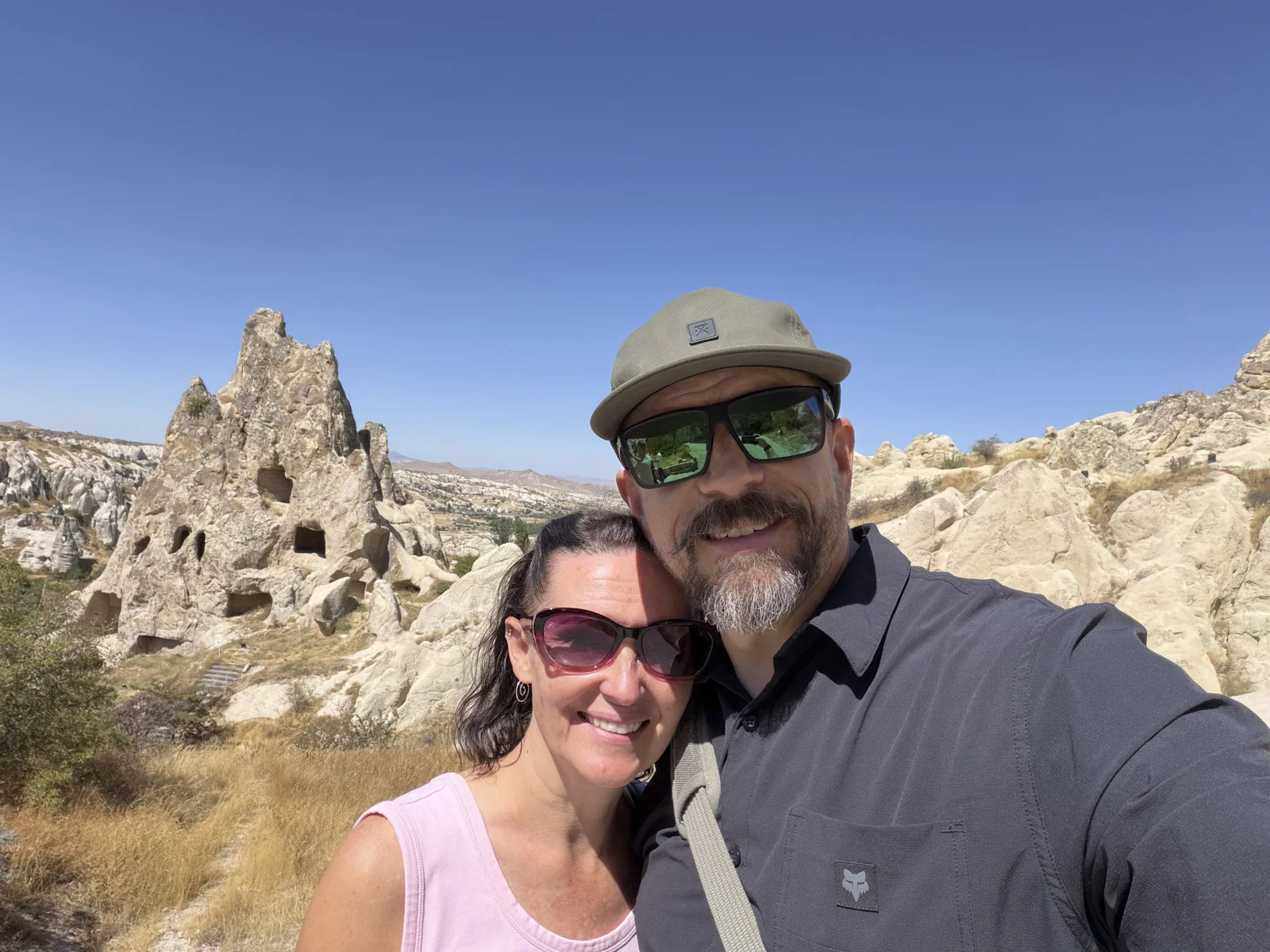 A couple taking a selfie in front of a pale rock cone riddled with cave openings, under a clear blue sky.