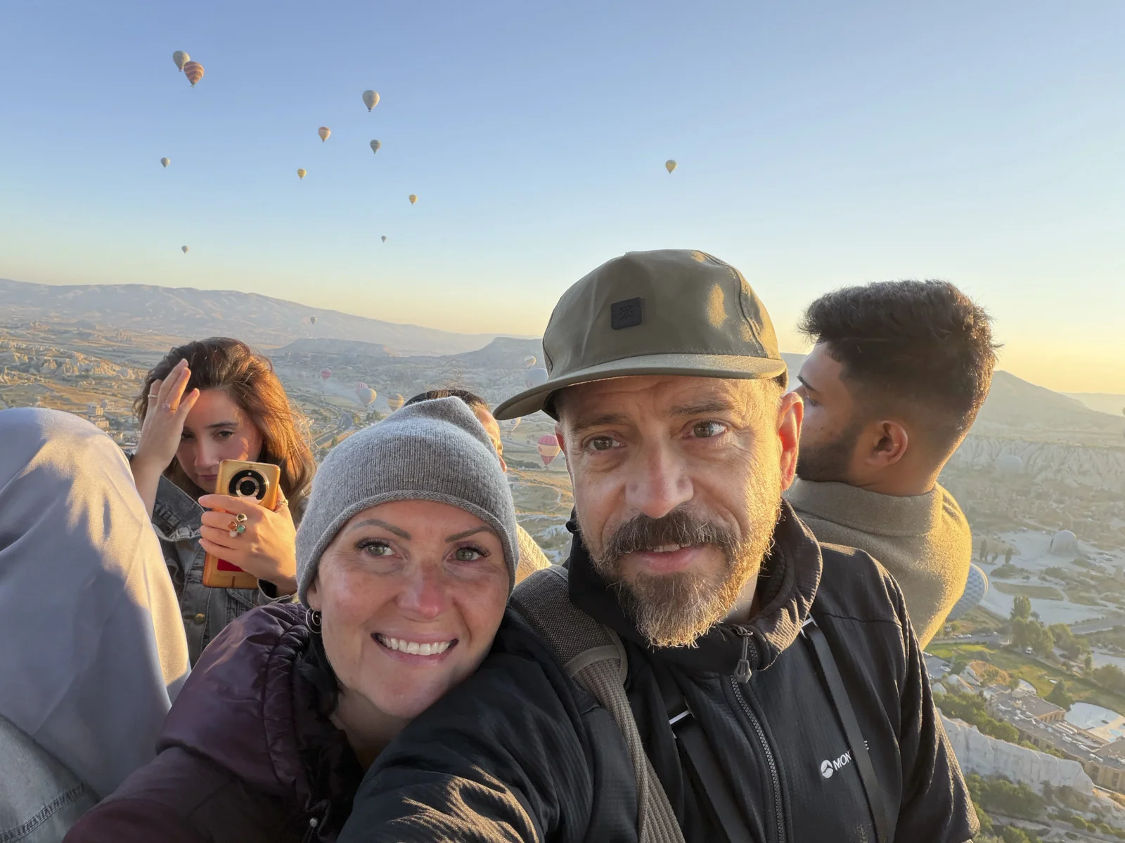 A group selfie in a hot-air balloon basket, with dozens of other balloons over the Cappadocian valleys at sunrise.