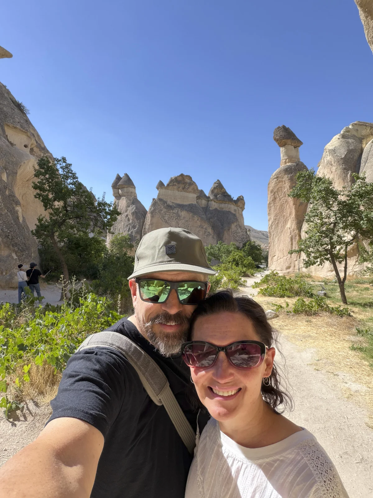 A couple taking a selfie on a dirt path among tall fairy chimneys and green trees under a blue sky.