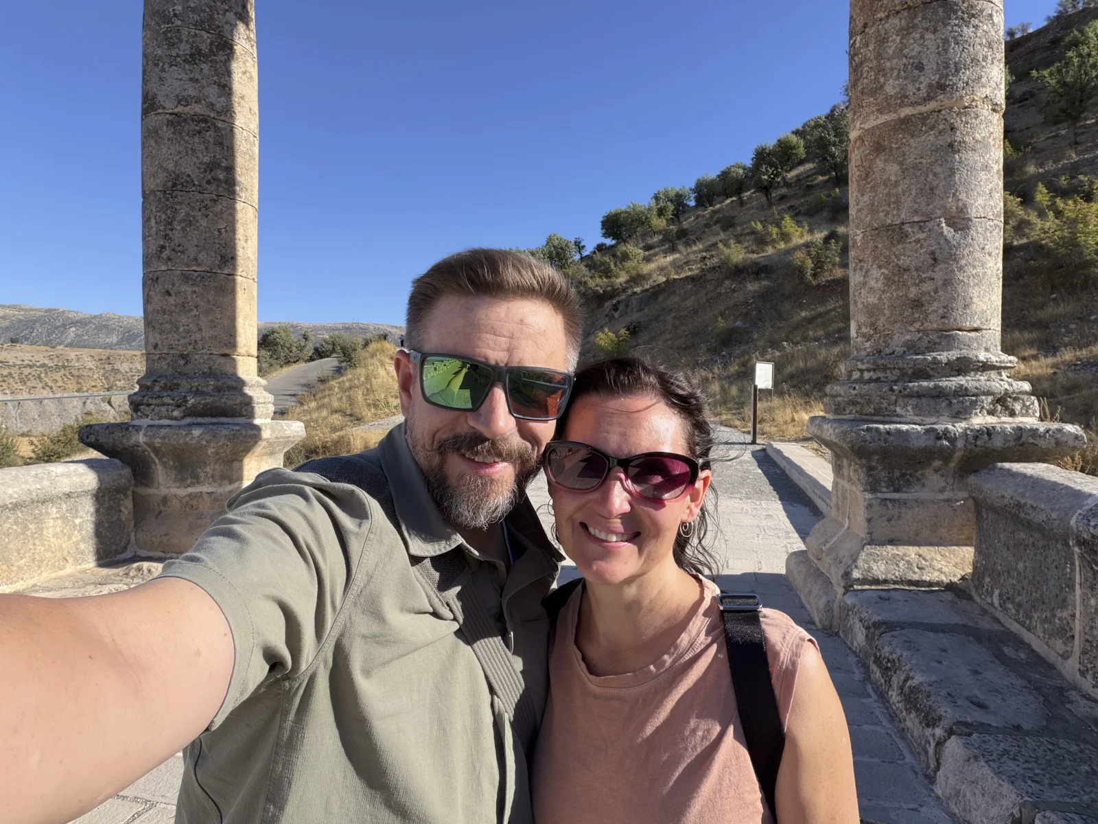 A couple taking a selfie between weathered stone columns of a Roman bridge, dry hills behind them.