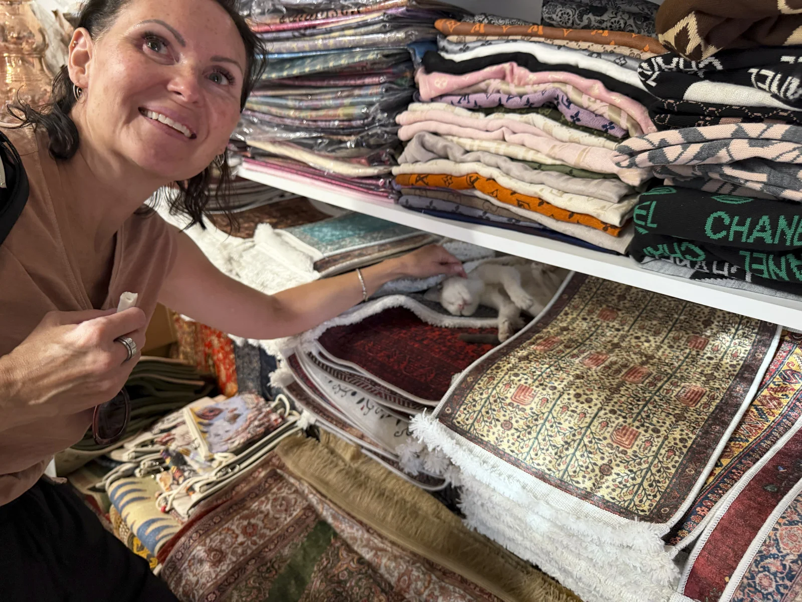 A woman reaching toward a small white cat asleep on folded rugs under a shelf of stacked textiles.