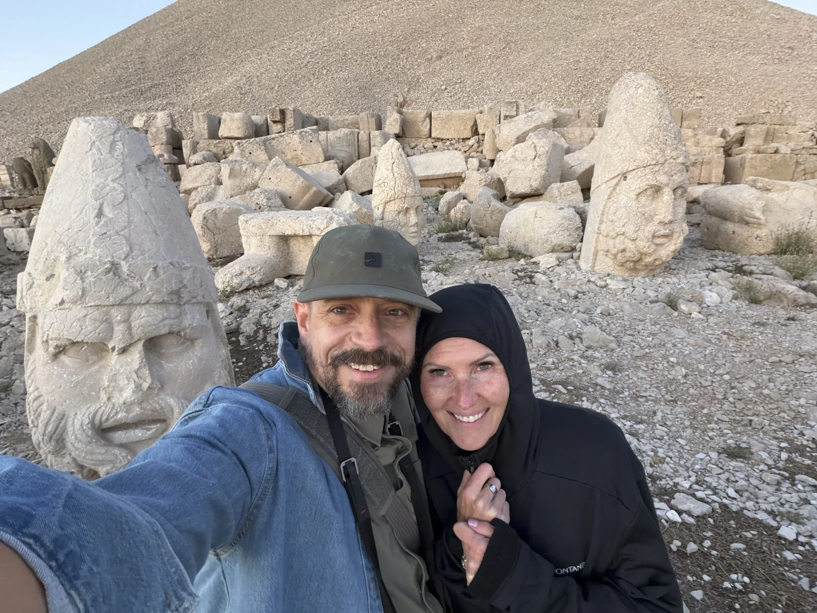A couple taking a selfie among colossal stone heads and rubble on the gravel summit of Mount Nemrut.