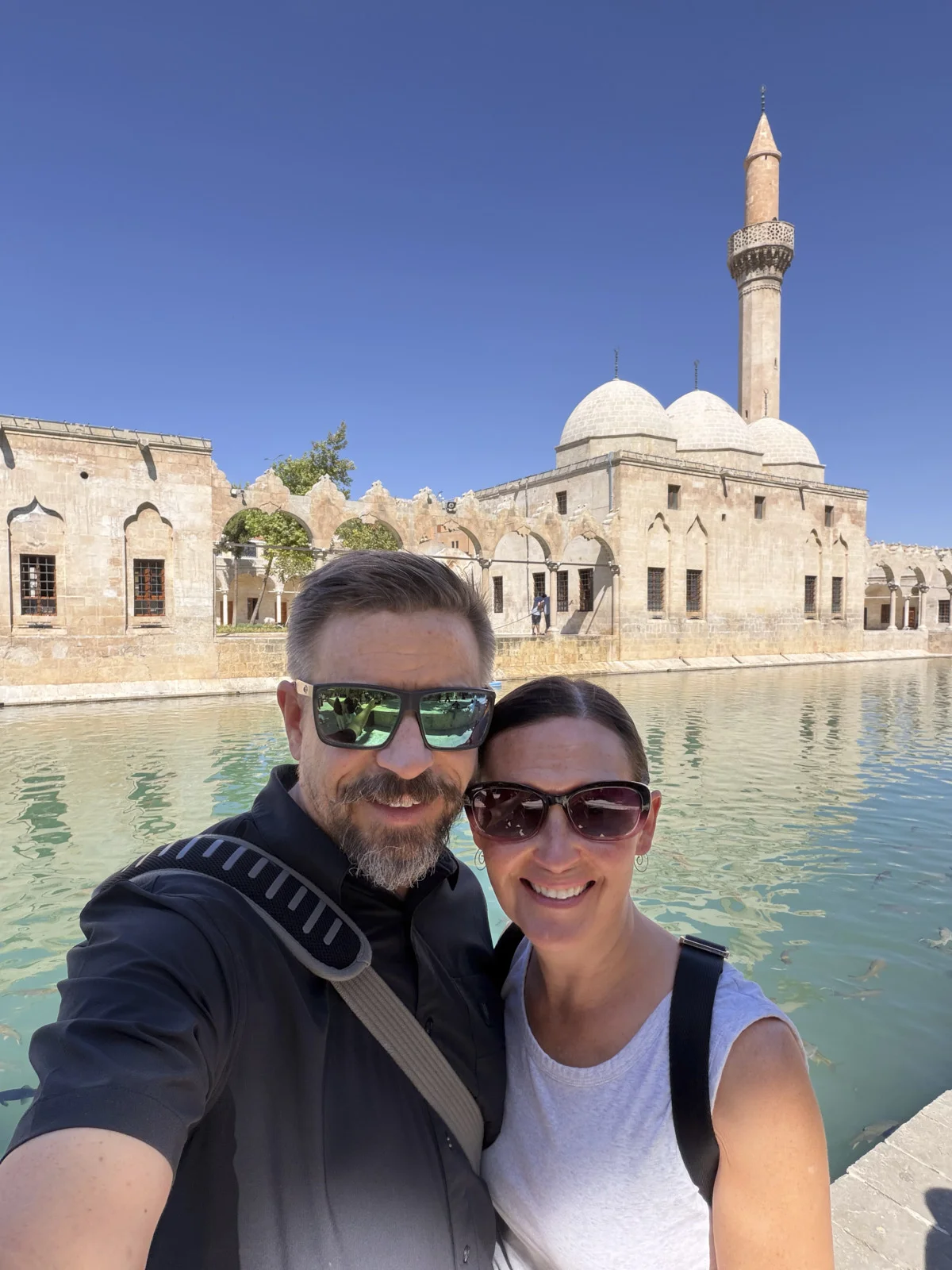A couple taking a selfie in front of a mosque with a minaret and a green pool full of fish.
