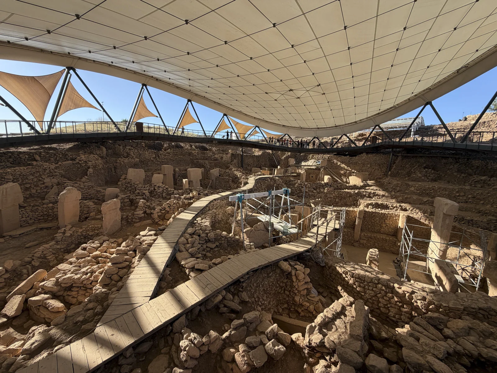 A wooden boardwalk curves through excavated stone circles under the broad white canopy at Göbekli Tepe.