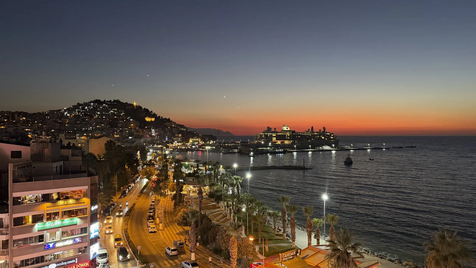 A lit cruise ship in a harbor at dusk, with a palm-lined waterfront road and a hillside of lights.