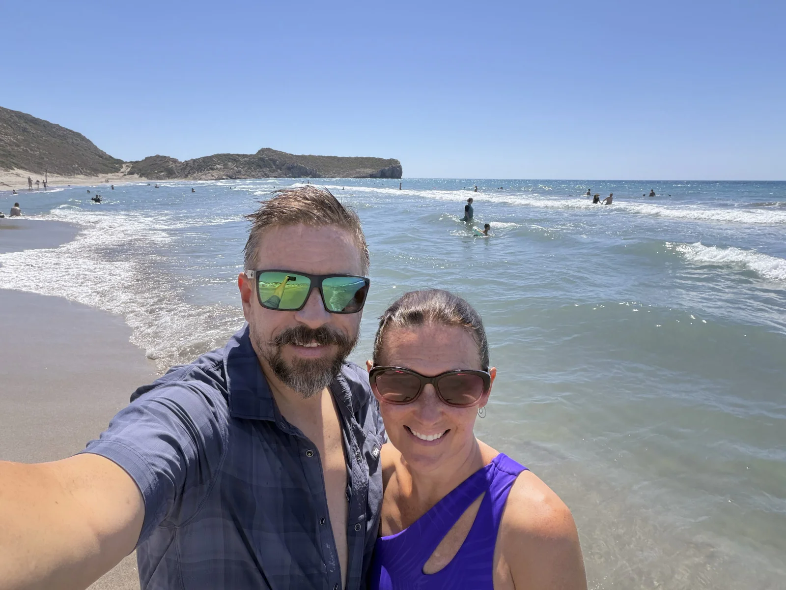 A couple taking a selfie at the edge of a wide sandy beach, with swimmers in the surf and a rocky headland.