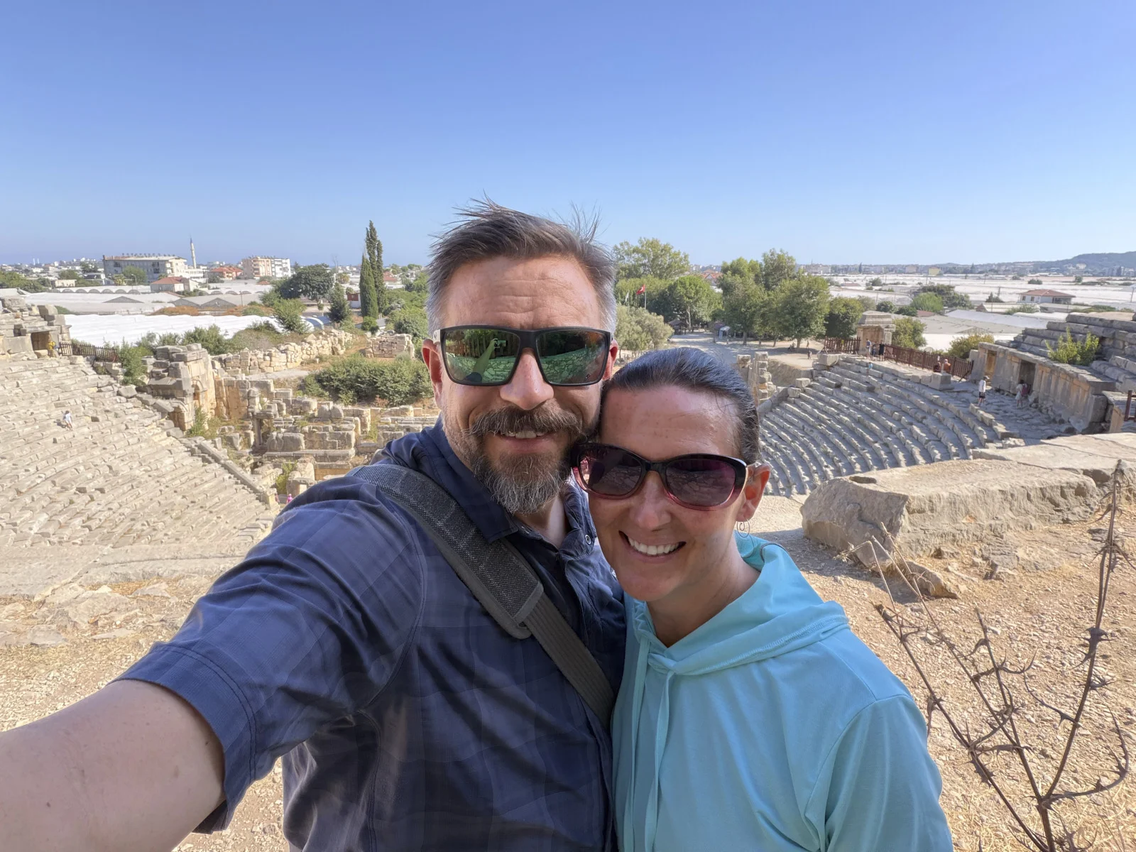 A couple taking a selfie above the curved stone seats of an ancient theater, with the town beyond.
