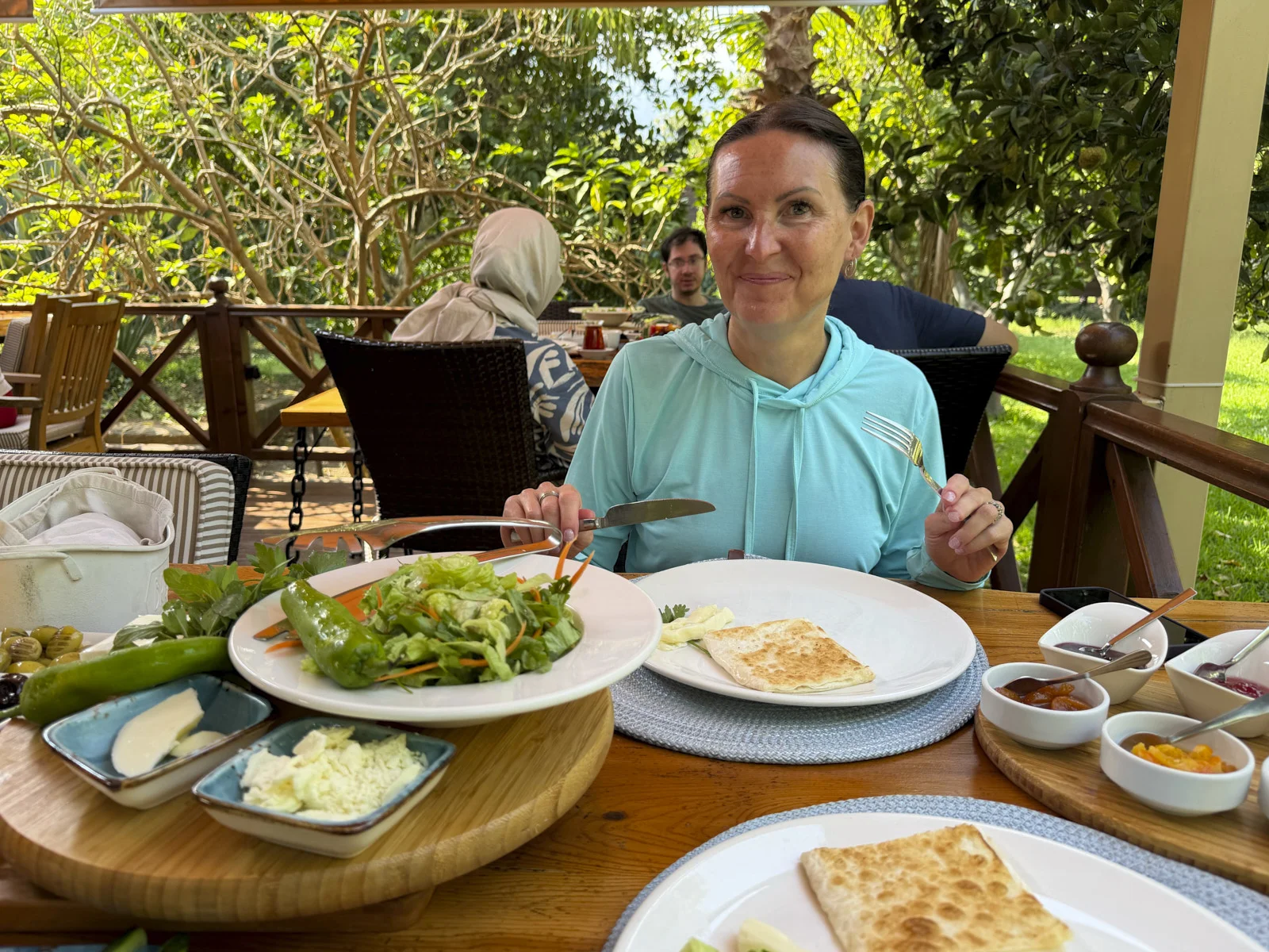A woman in a light blue hoodie at a breakfast table with gözleme, salad, cheese, and olives, in a shaded garden.