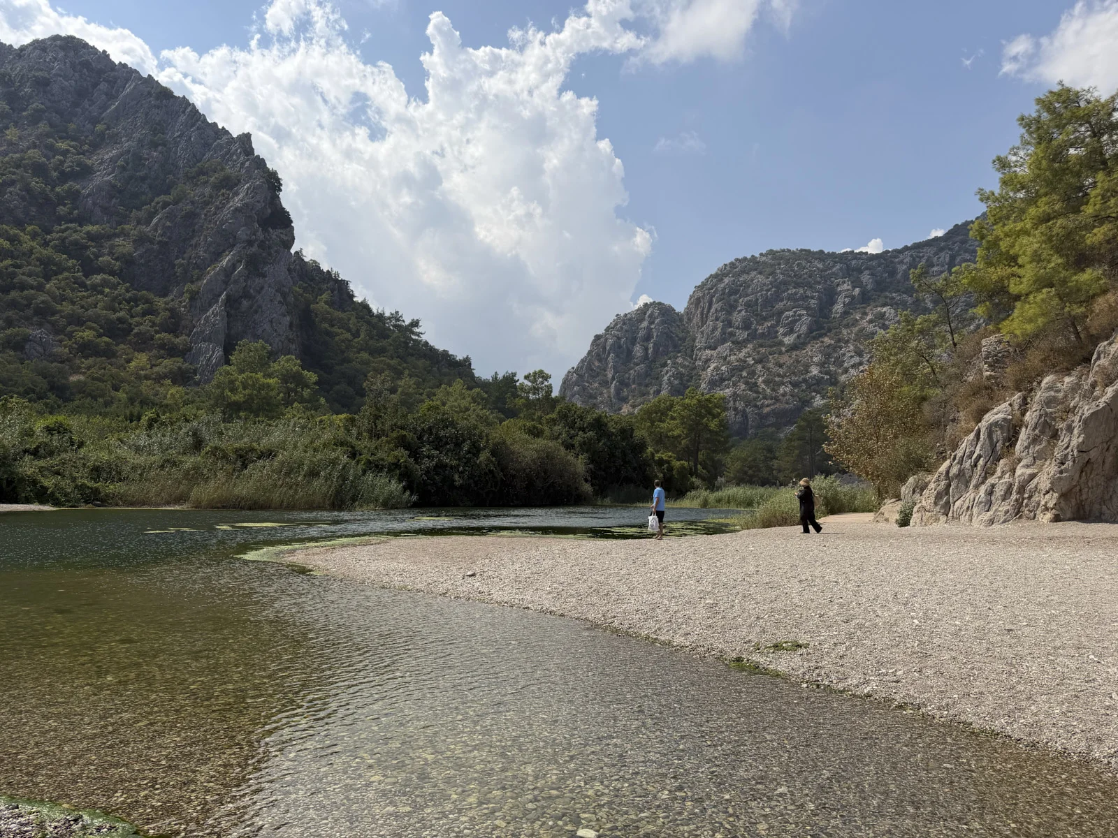 A clear shallow river and a gravel bar between steep green mountains, with two people on the far shore.