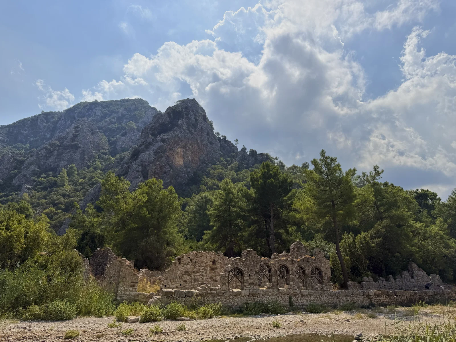 A ruined stone wall with arched openings at the foot of a steep forested mountain.