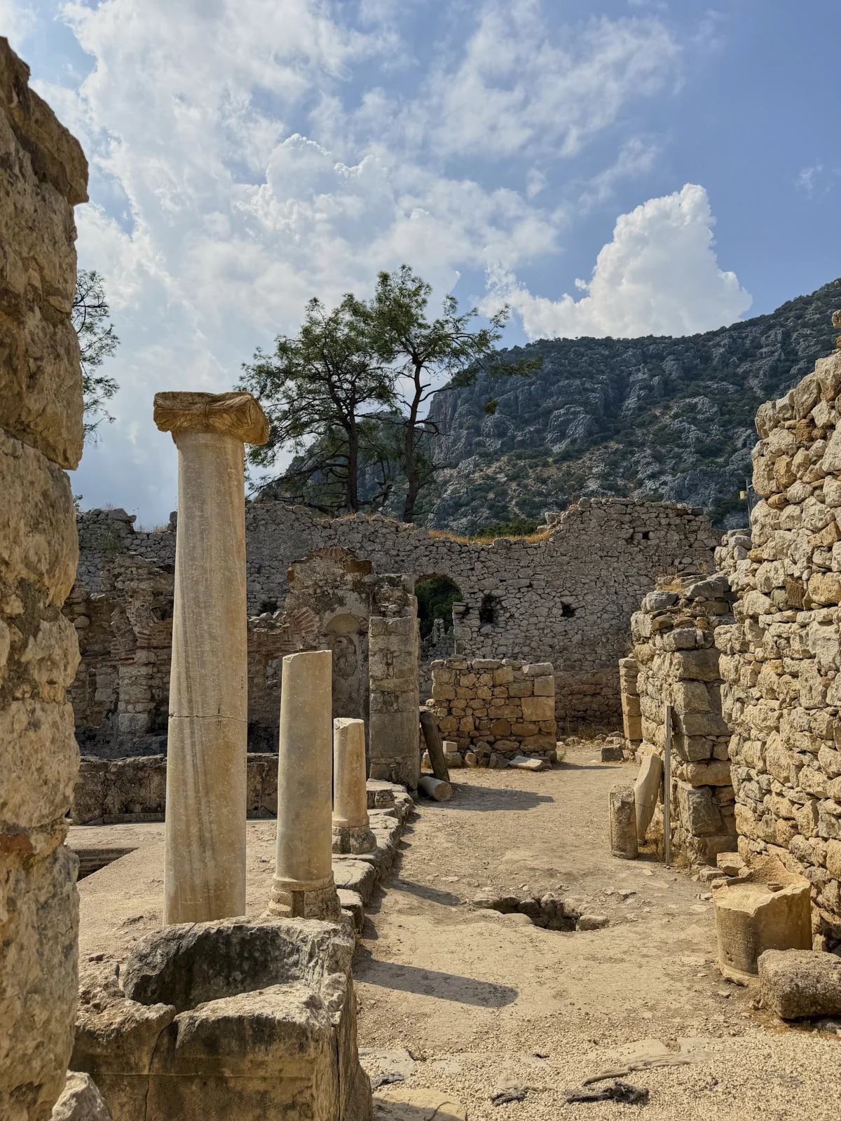 Fallen marble columns and broken walls inside an ancient ruin, with a pine and a mountain slope beyond.