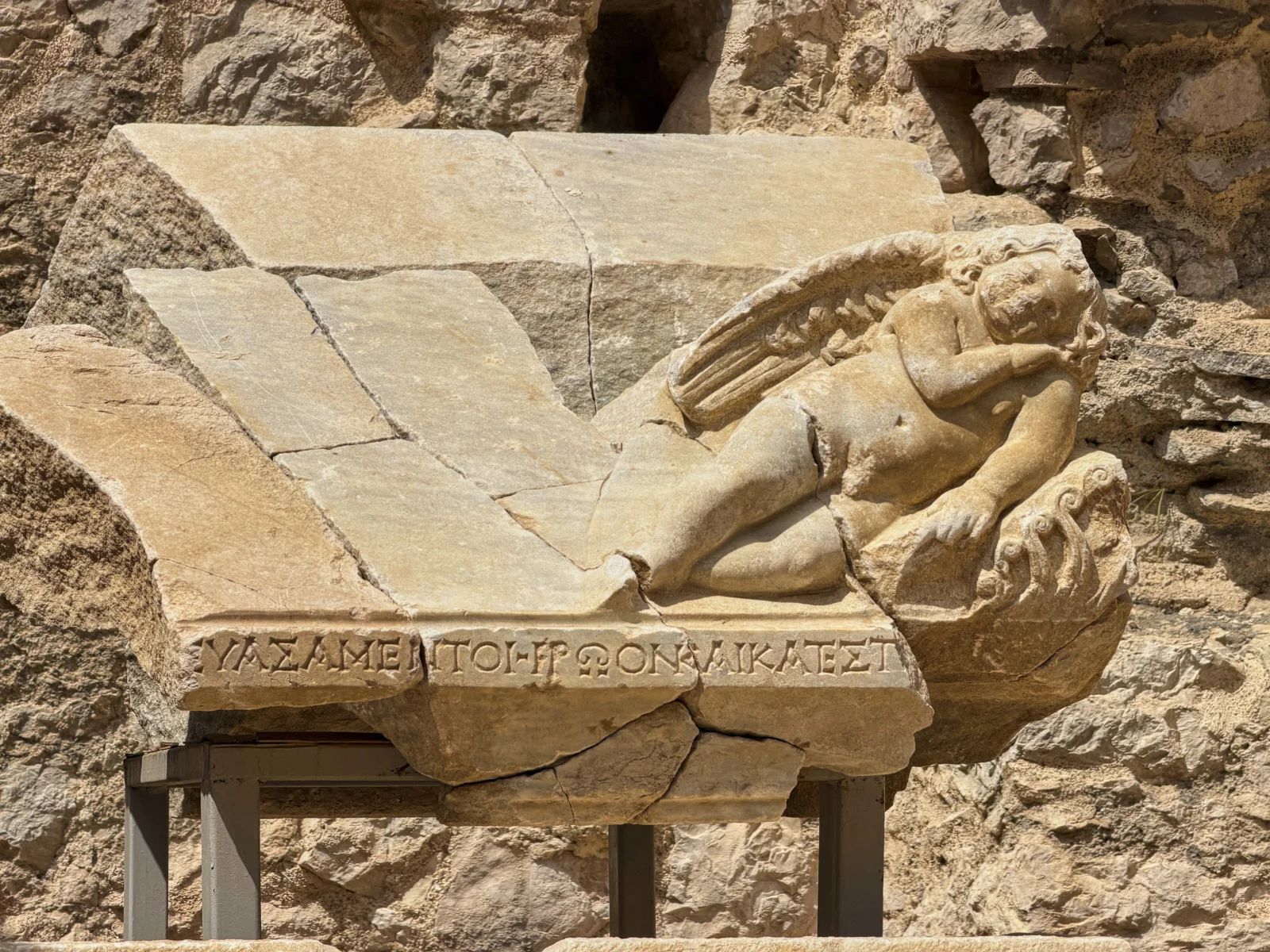 A marble relief of a winged child reclining on a broken sarcophagus lid, with a Greek inscription along the edge.