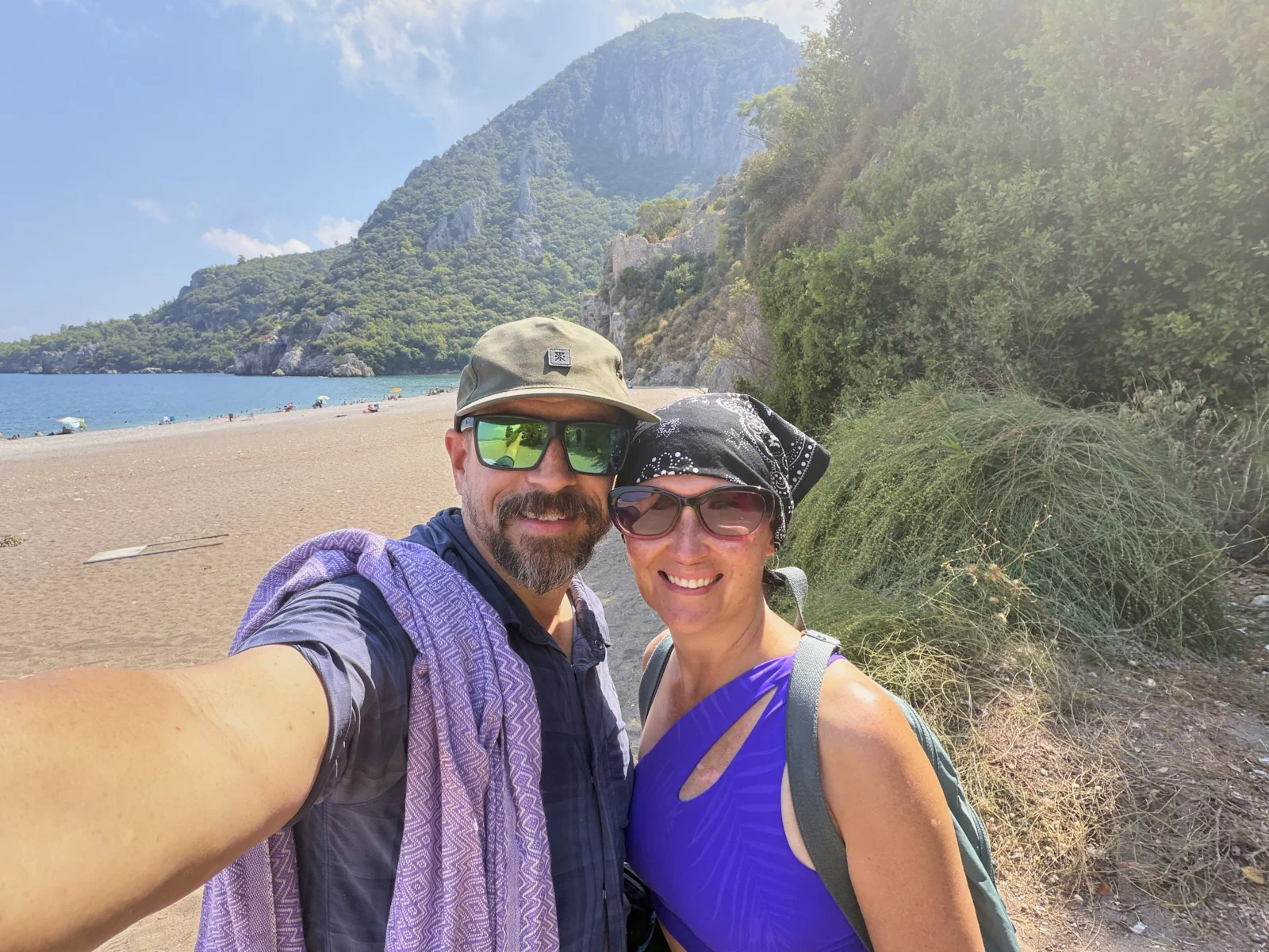 A couple taking a selfie on a pebble beach, with a steep green mountain and the sea behind them.